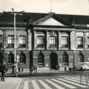 monumentalny, klasycystyczny budynek z kolumnowym wejściem i dekoracyjnym frontonem; przed budynkiem stoją stare samochody, a na chodniku przechodzą ludzie