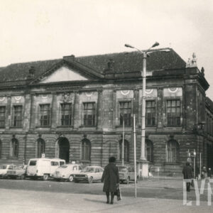 monumentalny, klasycystyczny budynek z kolumnowym wejściem i dekoracyjnym frontonem; przed budynkiem stoją stare samochody, a po chodniku przechodzą ludzie