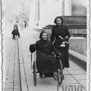 dwie młode kobiety idą chodnikiem, jedna z nich siedzi na wózku inwalidzkim, obie ubrane w ciemne płaszcze i nakrycia głowy