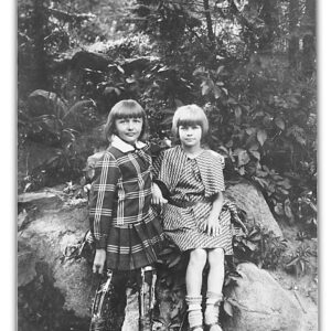 dwie małe dziewczynki w sukienkach siedzą na wielkim głazie, nogi jednej z nich znajdują się w metalowych szynach, za nimi gęste krzewy i drzewa liściaste