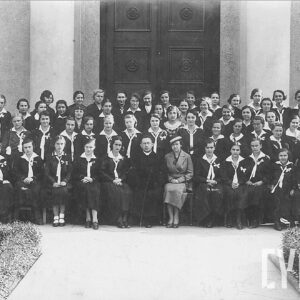 duża grupa dziewcząt ubranych w jednakowe mundurki i ustawiona w czterech rzędach (pierwszy rząd siedzi) na tle wielkich drzwi i fragmentu budynku, w pierwszym rzędzie siedzi ksiądz i jedna starsza kobieta, przed grupą fragment chodnika z trawnikami po obu stronach