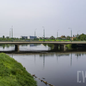 most łączący dwa brzegi, pod nim rzeka o wyraźnie podniesionym stanie; w głębi zarysy budynków
