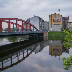 most łączący dwa brzegi, pod nim rzeka o wyraźnie podniesionym stanie; w głębi zarysy budynków