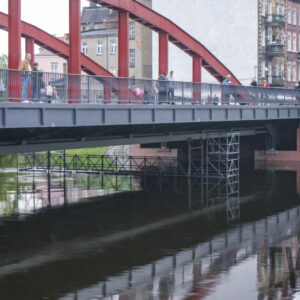 most łączący dwa brzegi, pod nim rzeka o wyraźnie podniesionym stanie; w głębi zarysy budynków