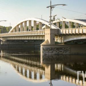 most łączący dwa brzegi, pod nim rzeka o wyraźnie podniesionym stanie; w głębi zarysy budynków