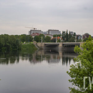 rzeka o wyraźnie podniesionym stanie, wzdłuż jej brzegów zielone, bujne drzewa, w głębi most, dalej dwa wysokie budynki