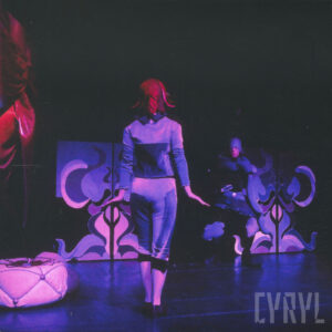 pośrodku sceny odwrócona tyłem kobieta, w głębi scenografia i schowany za nią mężczyzna w kostiumie