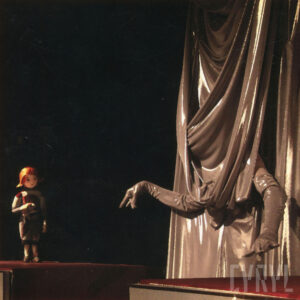 scena ze spektaklu, z lewej lalka, z prawej ludzkie ręce wychodzące z szarej, połyskującej tkaniny, stanowiące element scenografii