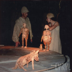 na pierwszym planie na okrągłej podwyższonej platformie trzy drewniane lalki: mężczyzny, kobiety i psa, w głębi w cieniu kobieta i mężczyzna