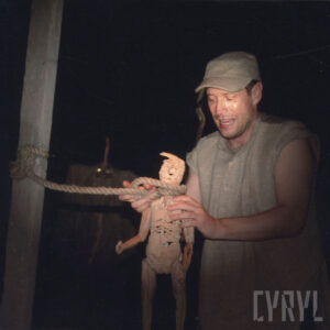 mężczyzna trzyma w rękach drewniana lalkę chłopca, która jest podwiązana sznurem do drewnianego słupa z lewej strony