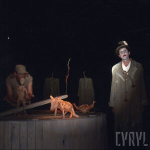 na pierwszym planie okrągła, drewniana platforma, na niej trzy drewniane lalki, w głębi z lewej mężczyzna trzyma w rękach lalkę chłopca, z prawej stoi mężczyzna w w cylindrze i płaszczu ze sznurem zawiązanym na szyi