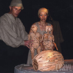 na pierwszym planie drewniana, okrągła platforma przy niej stoją kobieta i mężczyzna, którzy trzymają w rękach drewniane lalki kobiety i mężczyzny, na pierwszym planie leży mała drewniana beczka