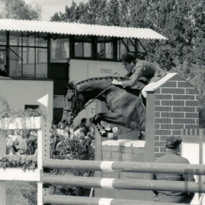 koń z jeźdźcem skacze przez przeszkodę, w głębi duża grupa widzów, za nimi murowany budynek z dużymi oknami w górnej partii