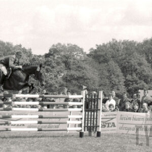 koń z jeźdźcem skacze przez przeszkodę, w głębi duża grupa widzów