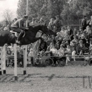 koń z jeźdźcem skacze przez przeszkodę, w głębi duża grupa widzów