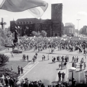 tłum ludzi z transparentami, w głębi okazały budynek z wieżą, z lewej dwa pomniki mężczyzny i krzyży