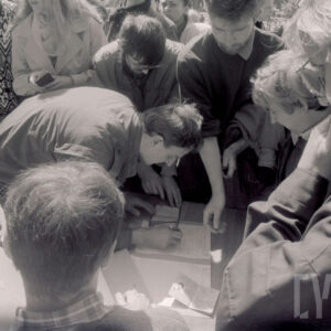 duża grupa ludzi pochylona nad stolikiem z dokumentami, jeden mężczyzna pisze