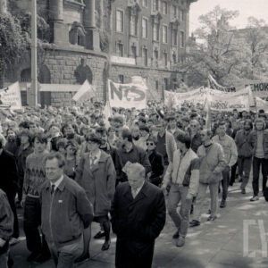 tłum ludzi z transparentami idzie ulicą, w głębi okazały gmach