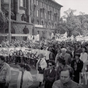 tłum ludzi z transparentami idzie ulicą, w głębi okazały gmach