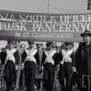 młodzi mężczyźni w historycznych mundurach maszerują w jednym szeregu, za nimi grupa oficerów w mundurach, nad głowami transparent z nazwą szkoły