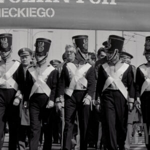 młodzi mężczyźni w historycznych mundurach maszerują w jednym szeregu, za nimi grupa oficerów w mundurach, nad głowami fragment transparentu z nazwą szkoły