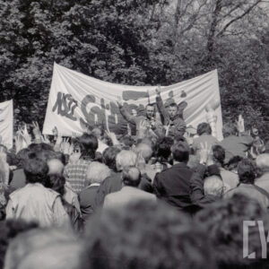 duża grupa ludzi stojąca tyłem do zdjęcia, w głębi dwóch mężczyzn stoi na podwyższeniu zwróceni twarzami do uczestników zgromadzenia