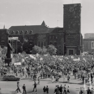 tłum ludzi z transparentami, w głębi okazały budynek z wieżą, z lewej dwa pomniki mężczyzny i krzyży