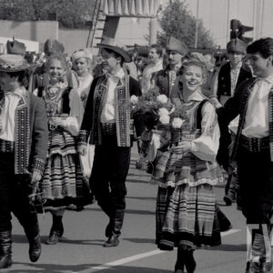 młodzi ludzie w strojach ludowych idą ulicą, część trzyma w rękach kwiaty, za nimi duża grupa ludzi