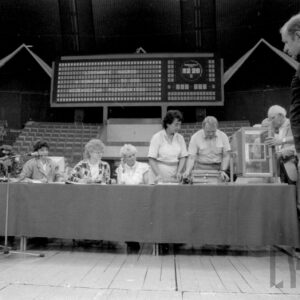 na pierwszym planie stół nakryty materiałem, przy nim siedzą ludzie, część osób stoi przy maszynie losującej, w głębi pusta widownia