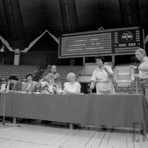 na pierwszym planie stół nakryty materiałem, przy nim siedzą ludzie, część osób stoi przy maszynie losującej, w głębi pusta widownia