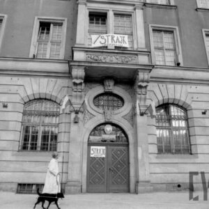 fragment okazałego budynku, pośrodku drzwi, na nich napis strajk, nad drzwiami rzeźbiony wykusz z oknem, przed budynkiem idzie kobieta z psem