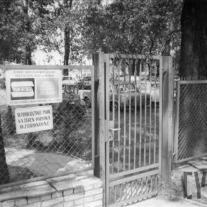metalowe ogrodzenie z furtką, z lewej tablice informacyjne, w głębi, za ogrodzeniem drzewa i zaparkowane samochody