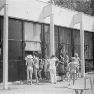 parterowy budynek z dużymi oknami, przy nim grupa ludzi stoi w kolejce przy okienku