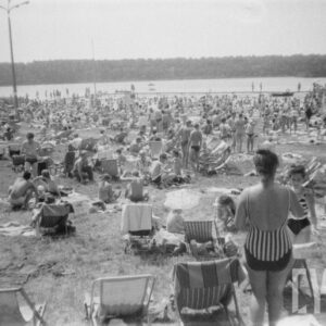 trawiasta plaża nad jeziorem, bardzo duża grupa ludzi odpoczywająca na leżakach i trawie, przy brzegu jeziora pomost, w głębi zalesiony brzeg