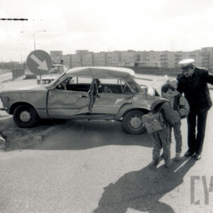 rozbity z prawej strony samochód osobowy, z prawej milicjant rozmawia z dójką chłopców i wskazuje im coś ręką, w głębi bloki mieszkalne