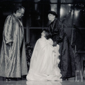 z lewej mężczyzna w długim szlafroku pochyla się nad klęczącą kobietą, z prawej kobieta w czarnej sukni trzyma dłoń na ramieniu klęczącej kobiety