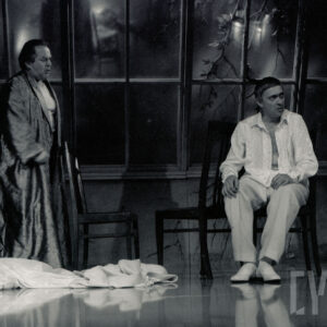 scena ze spektaklu, z lewej mężczyzna w szlafroku stoi, z prawej mężczyzna w jasnych spodniach i białej rozpiętej koszuli siedzi na krześle