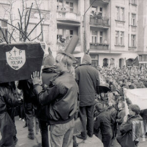 duże zgromadzenie osób z transparentami, z lewej grupa młodych osób stojąca na podwyższeniu trzyma na ramionach symboliczną trumnę, w głębi kilkupiętrowe kamienice