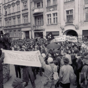 duże zgromadzenie osób z transparentami, z lewej grupa młodych osób stojąca na murku trzyma na ramionach symboliczną trumnę, część ma na głowach kaptury, w głębi kilkupiętrowe kamienice