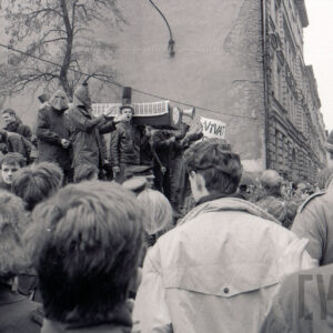 duże zgromadzenie osób z transparentami, w centrum grupa młodych osób stojąca na podwyższeniu trzyma na ramionach symboliczną trumnę, część z nich ma na głowach kaptury z wyciętymi otworami na oczy, w głębi ściana kamienicy bez okien