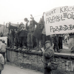 duża grupa młodych osób stojąca na murku, z lewej kilka osób trzyma na ramionach symboliczną trumnę, z prawej rozpięty transparent, w głębi murowane budynki przemysłowe