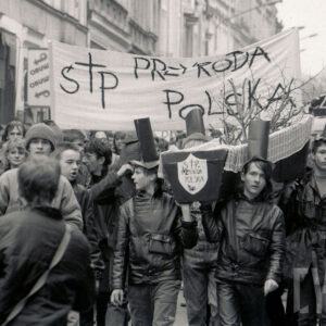 duże zgromadzenie młodych osób z transparentami, pośrodku młodzi chłopacy niosą na ramionach symboliczną trumnę, w głębi kilkupiętrowe kamienice