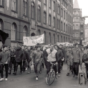 przemarsz młodych osób z transparentami, w głębi kamienice