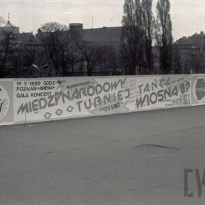 reklama zawieszona na barierce wzdłuż chodnika, chodnikiem z prawej idzie mężczyzna, w głębi korony drzew, za nimi zabudowania