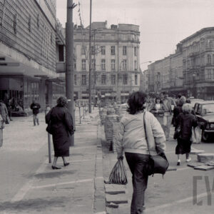 ruchliwa ulica, ludzie ze względu na remont chodnika idą po ulicy, wzdłuż ulicy stoją samochody osobowe, przy krawężnikach i na ulicy ułożone płyty chodnikowe, z lewej fragment budynku, w głębi widoczne skrzyżowanie i kilkupiętrowe kamienice