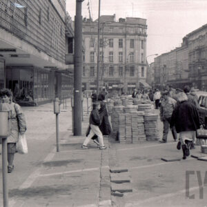 ruchliwa ulica, ludzie ze względu na remont chodnika idą po ulicy, wzdłuż ulicy stoją samochody osobowe, przy krawężnikach i na ulicy ułożone płyty chodnikowe, z lewej fragment budynku, w głębi widoczne skrzyżowanie i kilkupiętrowe kamienice