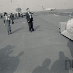 płyta lotniska, z prawej fragment schodów prowadzących do samolotu, na płycie stoją ludzie, w głębi samolot