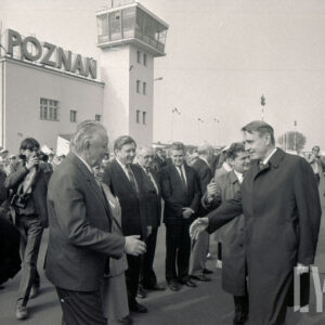 duża grupa ludzi zgromadzona na płycie lotniska, na pierwszym planie dwóch mężczyzn stojących twarzą do siebie z wyciągniętymi ku sobie prawymi dłońmi, w głębi jasny budynek z wieżą i napisem Poznań