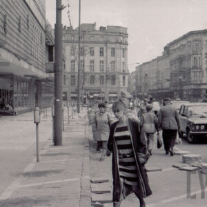 ruchliwa ulica, ludzie ze względu na remont chodnika idą po ulicy, wzdłuż ulicy stoją samochody osobowe, przy krawężnikach i na ulicy ułożone płyty chodnikowe, z lewej fragment budynku, w głębi widoczne skrzyżowanie i kilkupiętrowe kamienice