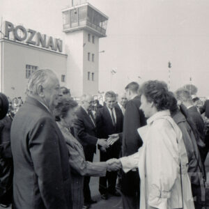 duża grupa ludzi zgromadzona na płycie lotniska, pośrodku dwie kobiety witają się, w głębi jasny budynek z wieżą i napisem Poznań